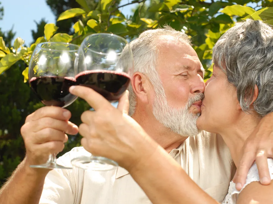 two retired couple kissing while holding red wine with green leaves at the background