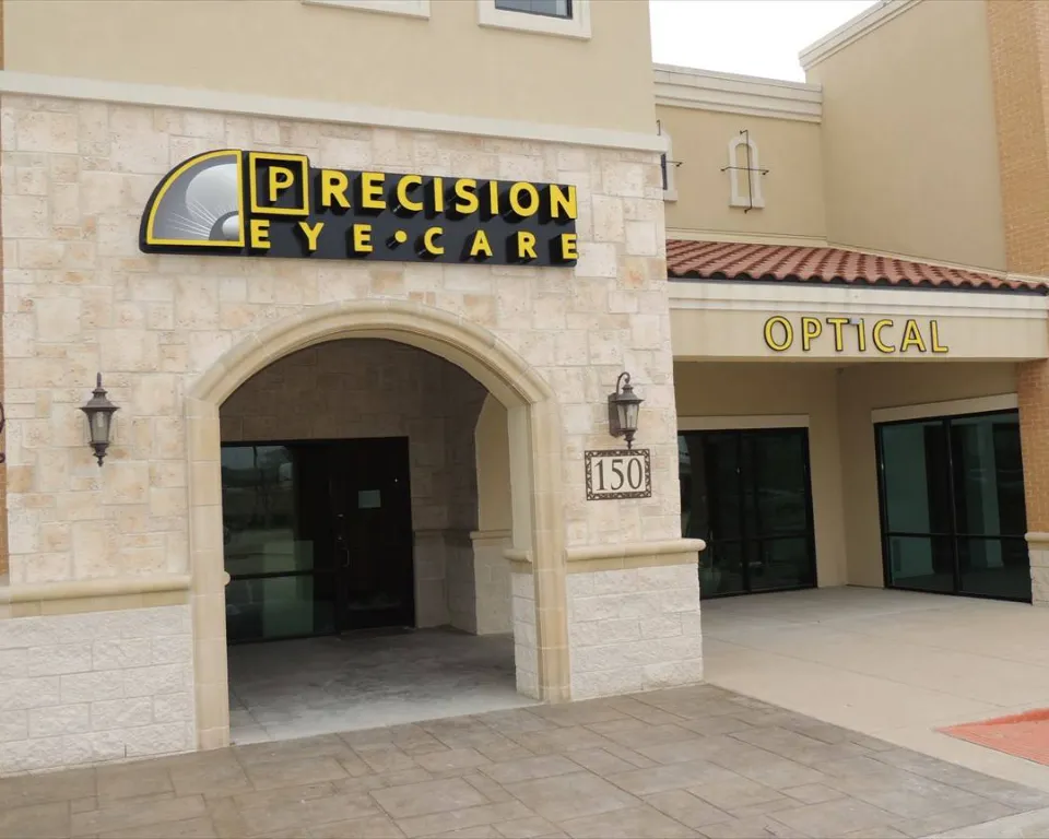 Exterior view of Precision Eye Care optical shop with glass doors and windows, a black arched entrance, and a yellow sign with the words 'Precision Eye Care' on a beige wall.