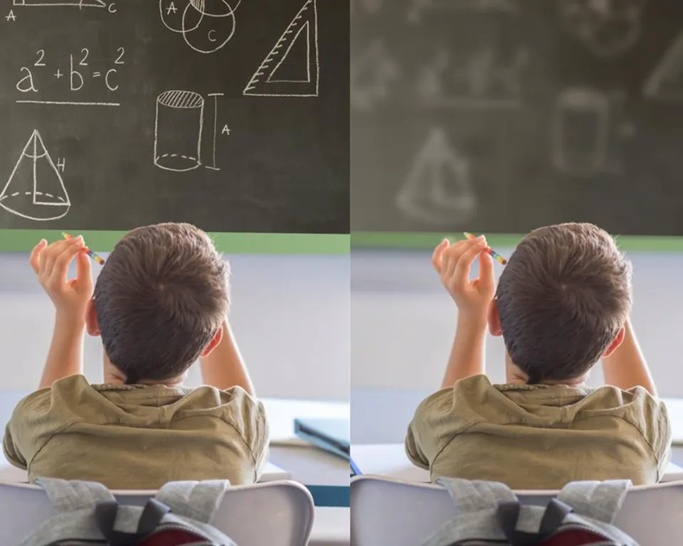 A young boy sitting in a classroom is looking at a blackboard with mathematical equations and shapes written on it.