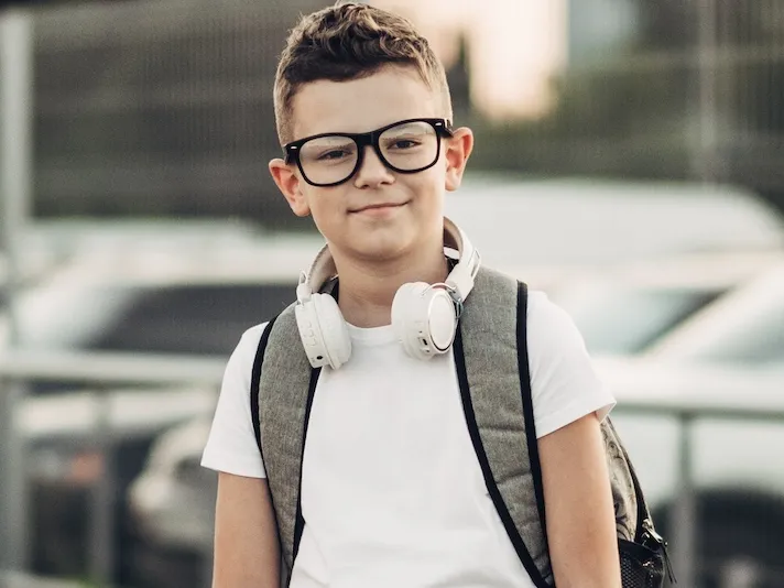 A boy wearing glasses, a white shirt, and a backpack with headphones is standing in front of a parking lot.
