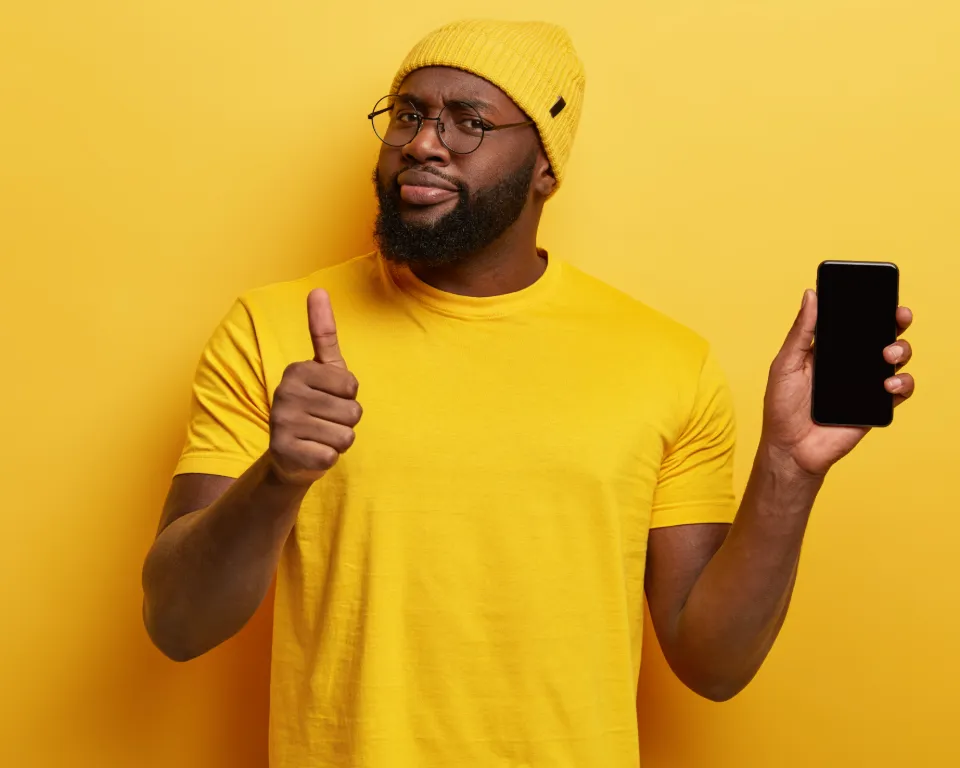 A man wearing a yellow shirt and hat is holding a smartphone and giving a thumbs-up gesture