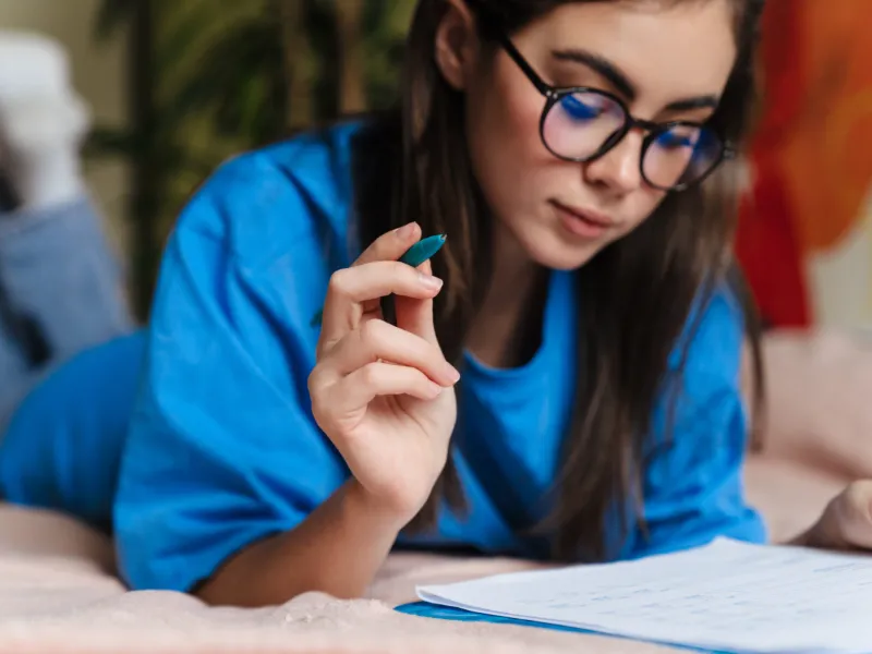 A young girl with glasses is reading a book on a bed and holding a pen.
