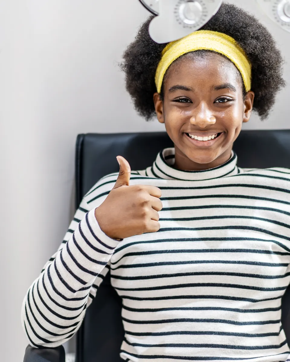 A smiling girl sitting in a chair wearing a yellow headband and a striped shirt