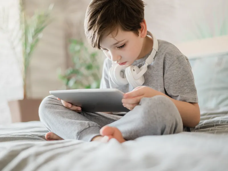 A boy with headphones sitting on a bed holding a tablet
