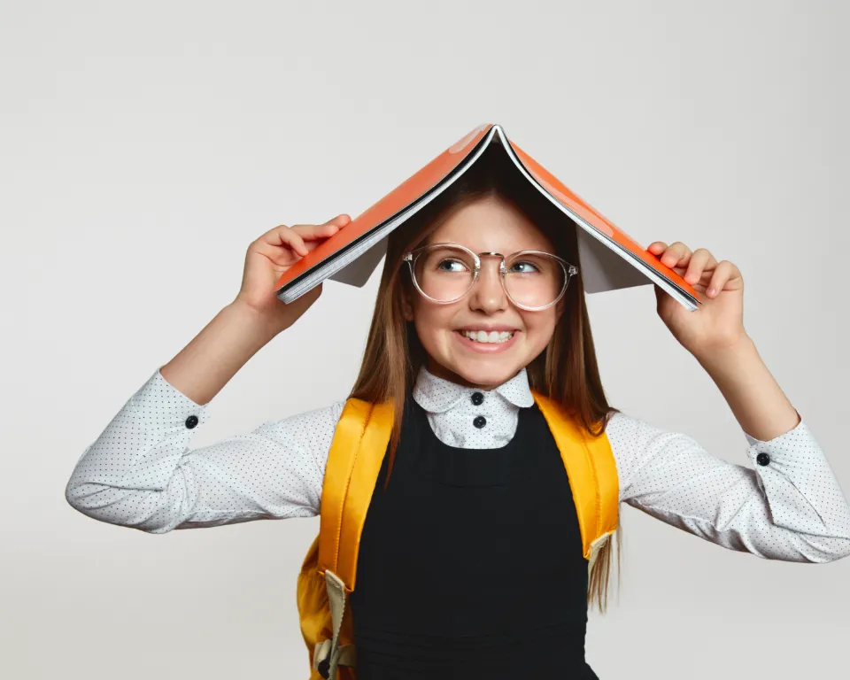 Girl holding a book over her head smiling and wearing glasses and a backpack