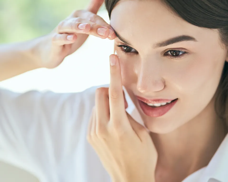 A woman with brown hair wearing a white shirt is applying makeup to her eyes while smiling and looking down. She is holding her eyebrows with her right hand and her left hand is on her cheek.