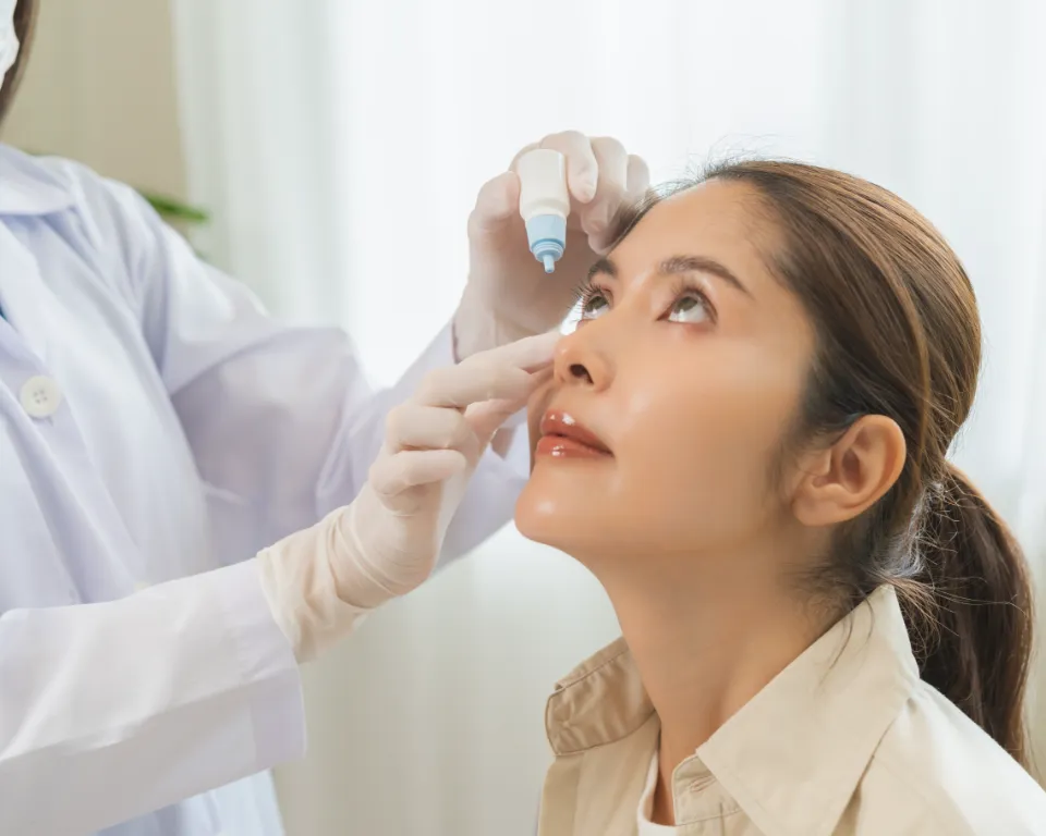 A doctor in a lab coat is administering eye drops to a woman in a hospital room.