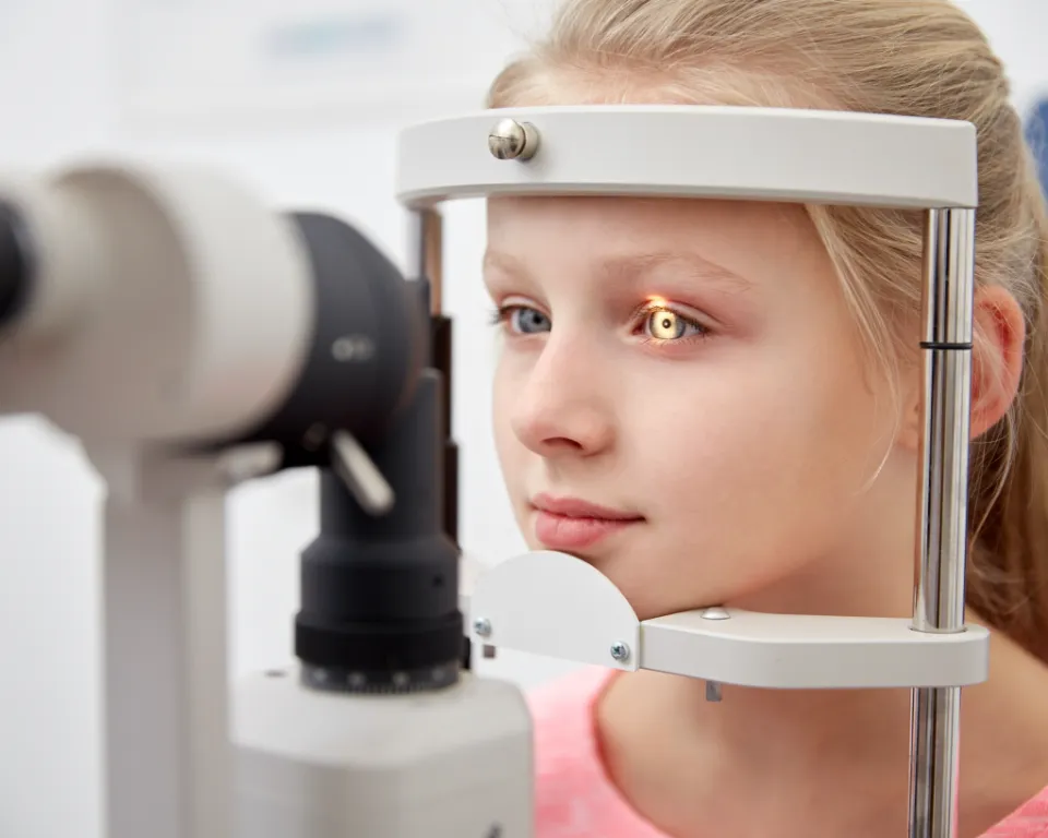 A young girl wearing a pink shirt is having her eyes examined with a slit lamp in an eye clinic