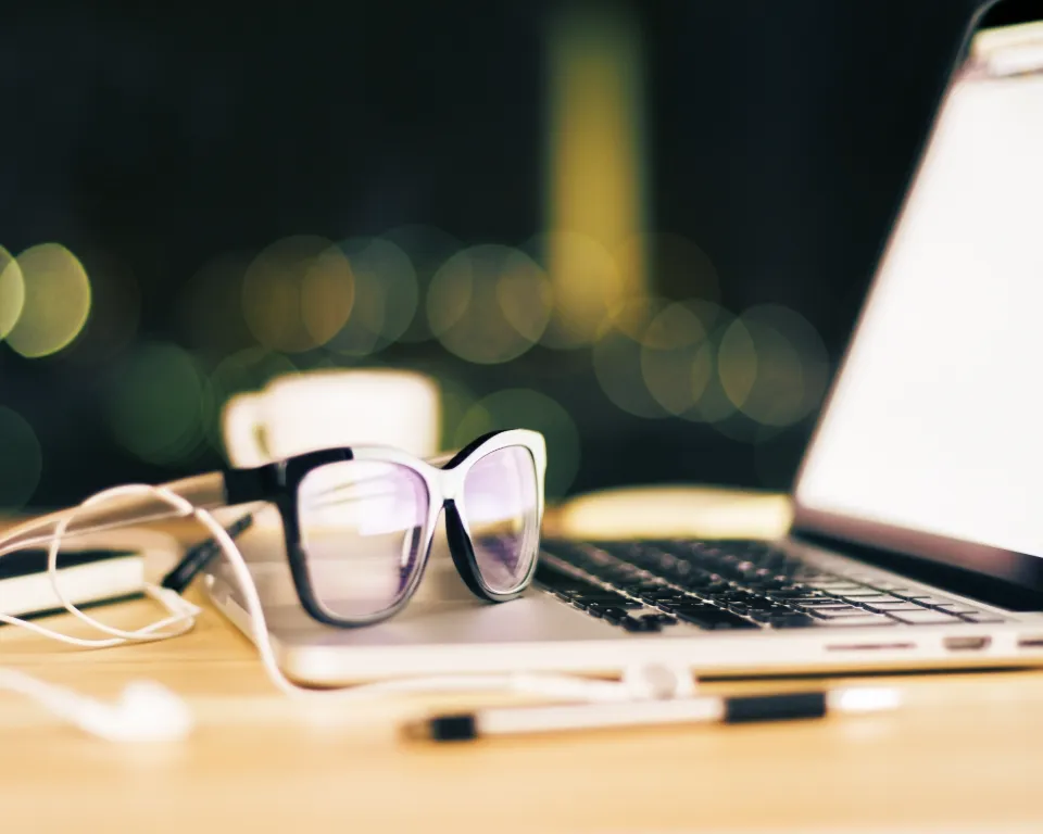 A laptop with a pair of sunglasses and headphones on a wooden table with a cup and a pen