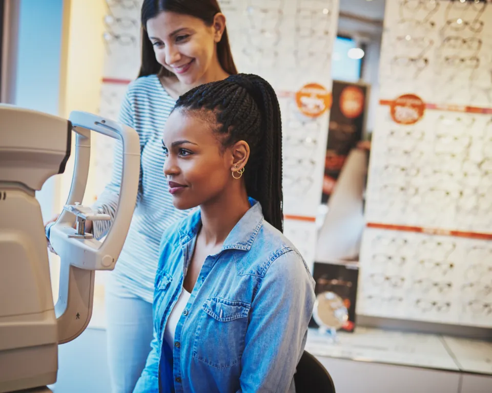 A woman is using an eye examination machine while another woman is smiling behind her, probably in an optical shop.