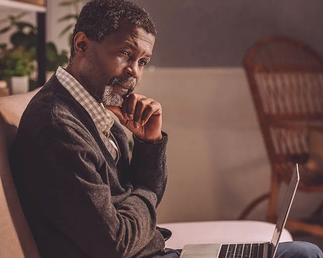 Thoughtful man sitting with open laptop on couch at home