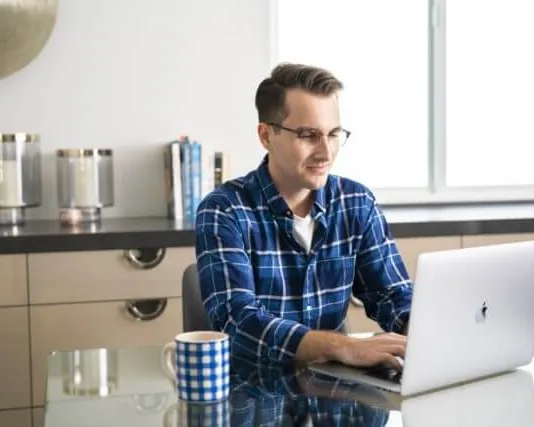 Man working on laptop at kitchen table with coffee mug nearby